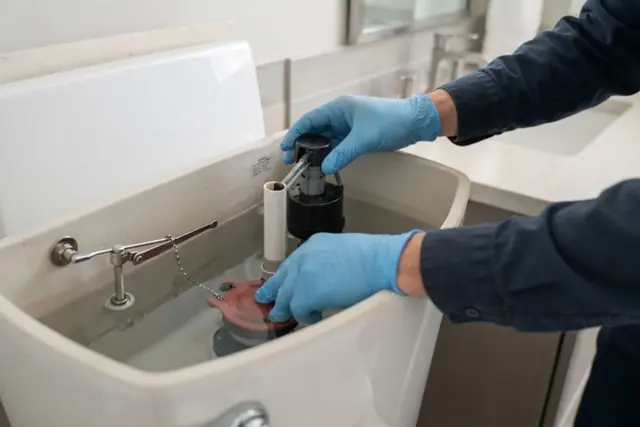 Gloved hands adjusting the flapper and fill valve inside an open toilet tank